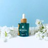 Flacon dropper bleu turquoise de parfum de lessive Douce Innocence avec bouchon en bois, entouré de fleurs blanches.